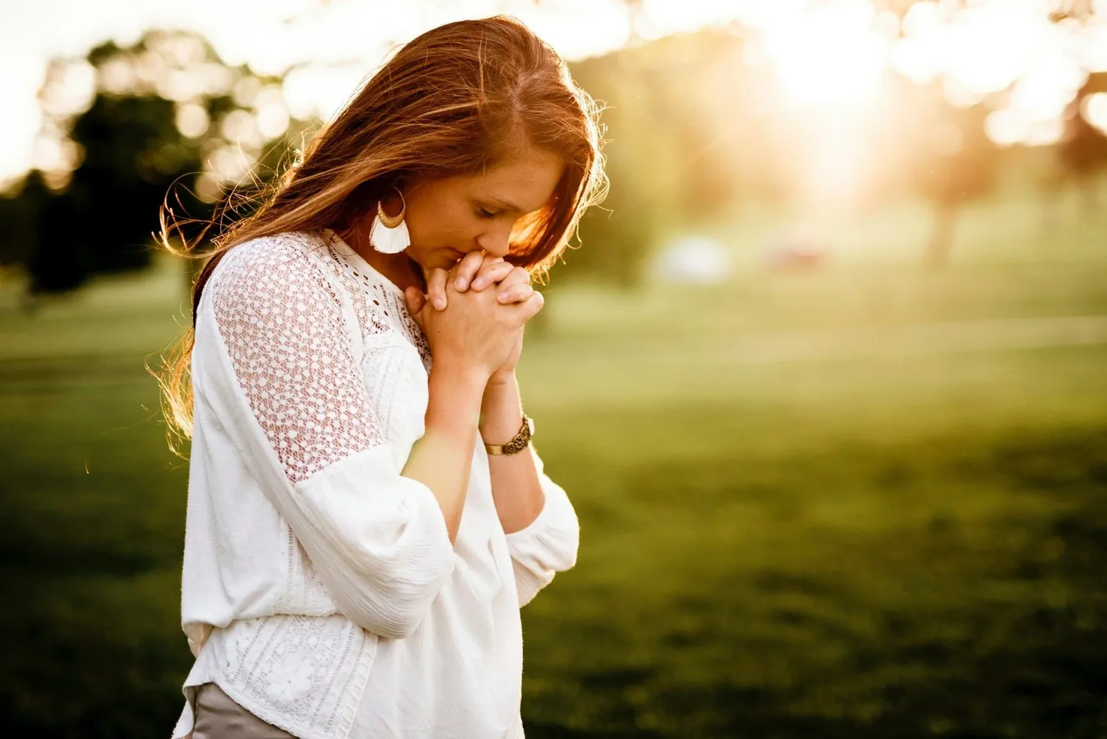 A woman standing outdoors in warm sunlight, eyes closed, hands held together